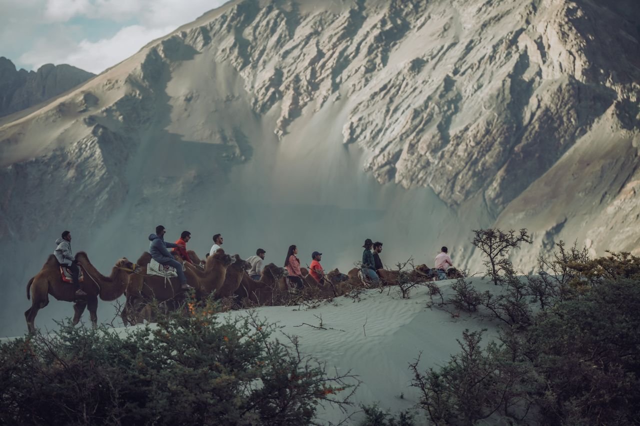 A group of tourists riding camels against a stunning mountain backdrop in daylight.